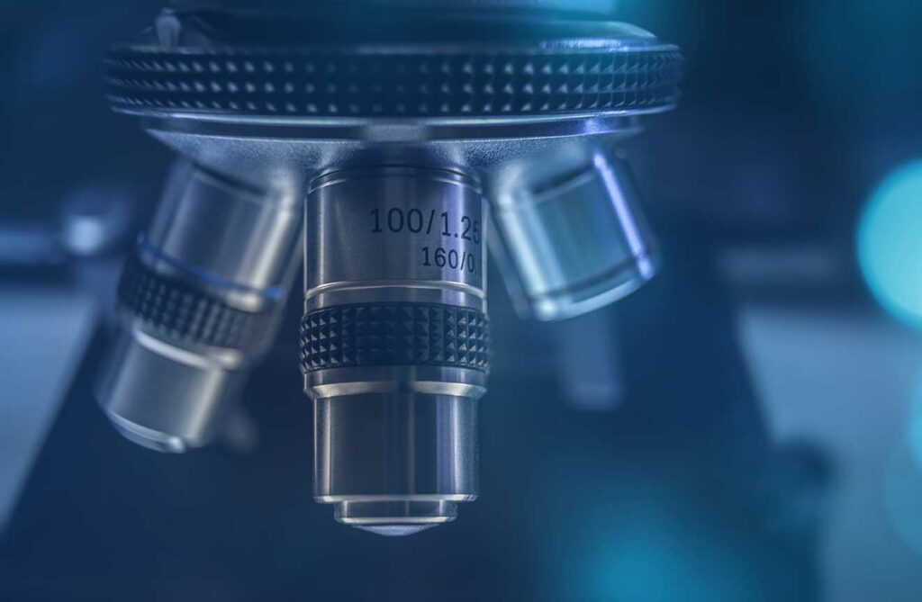 Close-up of a microscope’s 3 lenses, in focus against a blurred blue-lit background in medical lab setting